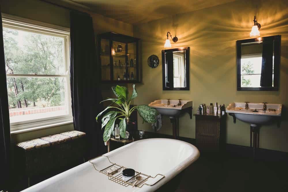 Interior view of bathroom with black mirrors over two Victorian wash stands, sash window and roll top bath with brass bath caddy.